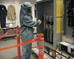 Trabajador con traje de protección contra arco inspeccionando un tablero, con el área delimitada por conos y cinta
